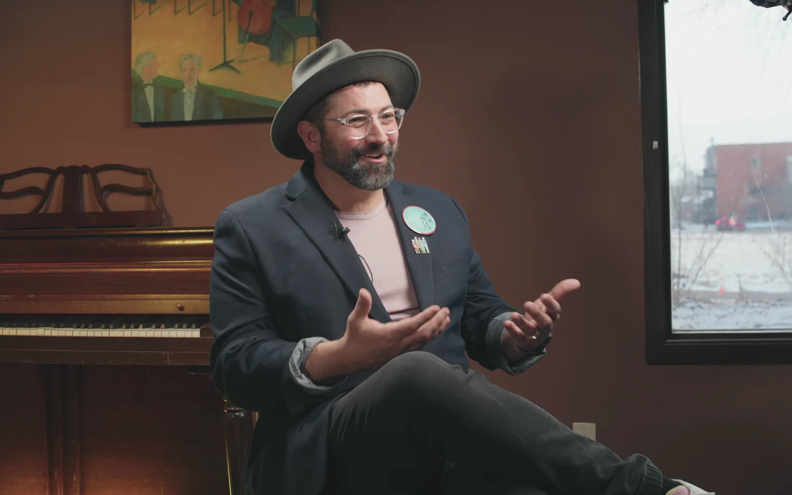 A man in a hat speaks while seated beside a piano.