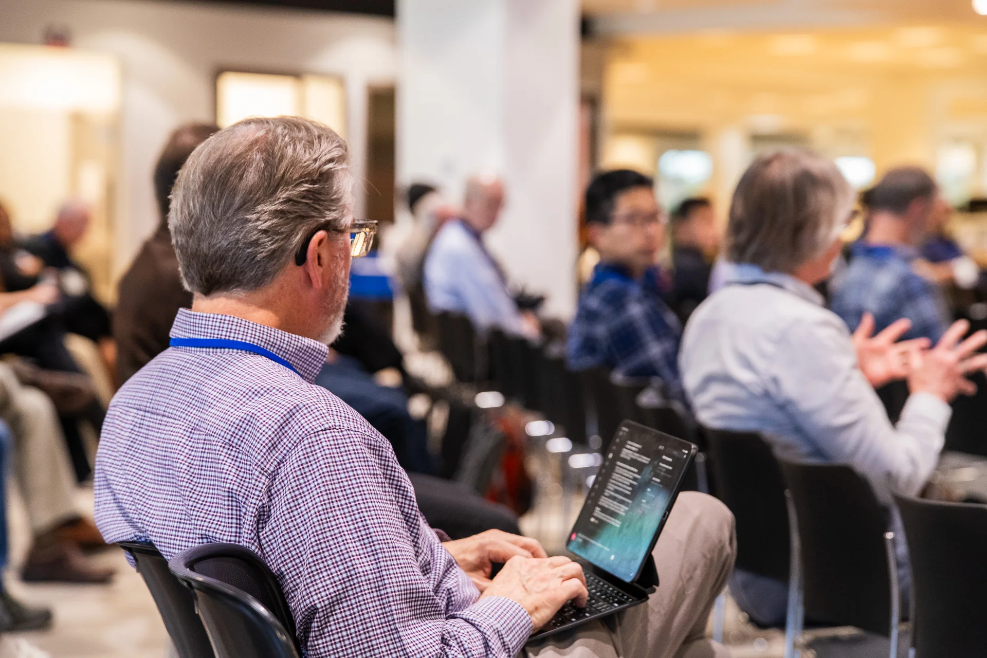 A seated conference attendee uses a laptop while listening among the audience.